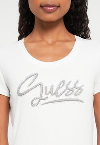 Frau trägt ein weißes T-Shirt mit silberne Strass-verziertem "Guess"-Logo auf der Brust und hat lockige, dunkle Haare, die auf den Schultern sichtbar sind.