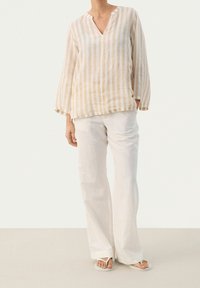 Blusa a righe in lino beige e crema con scollo a V, maniche lunghe a campana, abbinata a pantaloni bianchi a gamba larga e sandali semplici.
