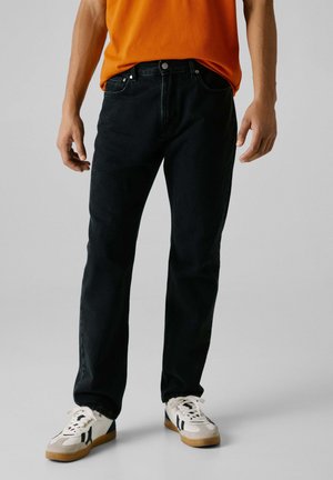 Pantalones vaqueros de mezclilla negra con un corte recto, cinco bolsillos y detalles de herrajes metálicos. Llevados con una camiseta naranja brillante y zapatillas blancas.