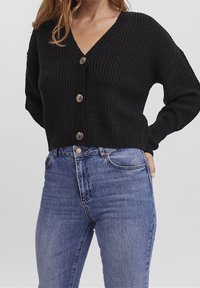 Zwarte gebreide cardigan met een v-hals, voorzien van vier grote knopen aan de voorkant. Gecombineerd met blauwe high-waisted jeans.