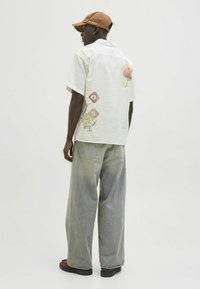 Short-sleeve, light-colored shirt with botanical graphics on the back, paired with loose, faded gray jeans and a brown cap.