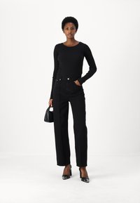 Vrouw in een zwart ribbeltrui met lange mouwen en een hoge taille, met zwarte broek, die een kleine zwarte handtas vasthoudt, gekleed in zwarte hakken, tegen een witte achtergrond.