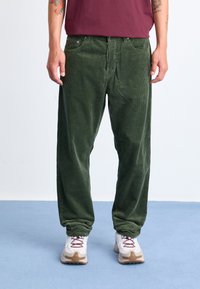 Green corduroy pants with a straight-leg design, featuring a textured surface and front pockets, paired with white and beige sneakers.