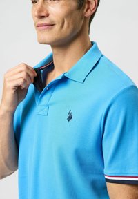 Lyseblå polo skjorte med klassisk krave, korte ærmer og stribede detaljer i rød og marineblå. Har et lille broderet logo på brystet.
