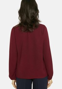 Pruunikas pikkade varrukatega särk, sile kangas, lõtv lõige, ümar serv, peene tekstuuriga, kantud tumesiniste pükstega, vaadatuna tagantpoolt.