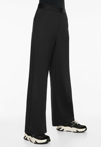 Wide-leg black trousers made of smooth fabric, featuring a sleek waistband and front pockets, paired with black and white chunky sneakers.