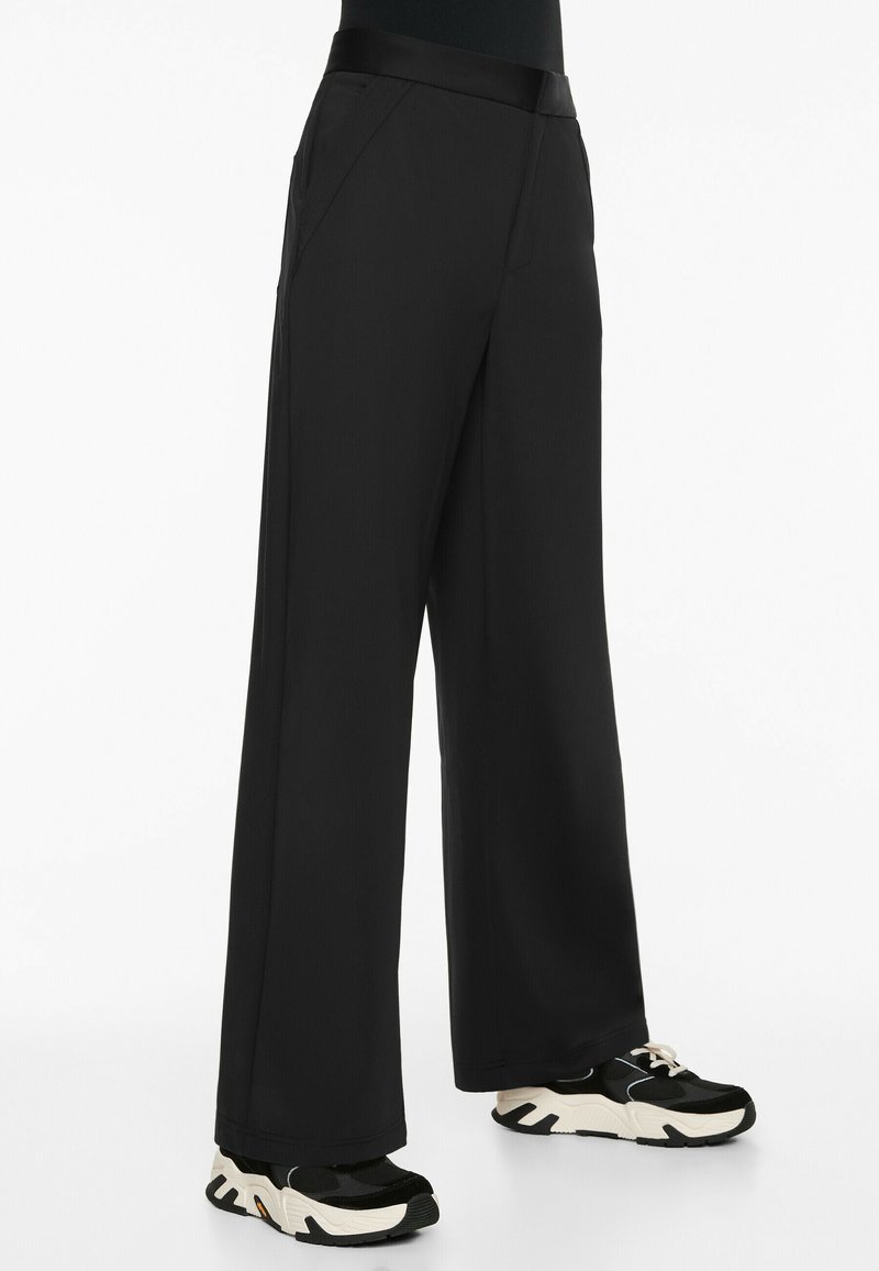 Wide-leg black trousers made of smooth fabric, featuring a sleek waistband and front pockets, paired with black and white chunky sneakers.