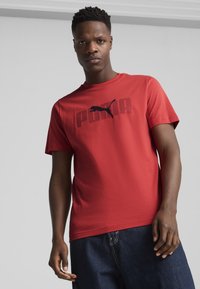 Rød bomulds t-shirt med korte ærmer, prydet med et sort Puma-logo og tekst over brystet. Glat tekstur, afslappet pasform.
