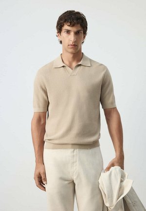 Joven con una camiseta tipo polo beige de manga corta texturizada y pantalones crema, sosteniendo una chaqueta de color claro en su mano derecha.