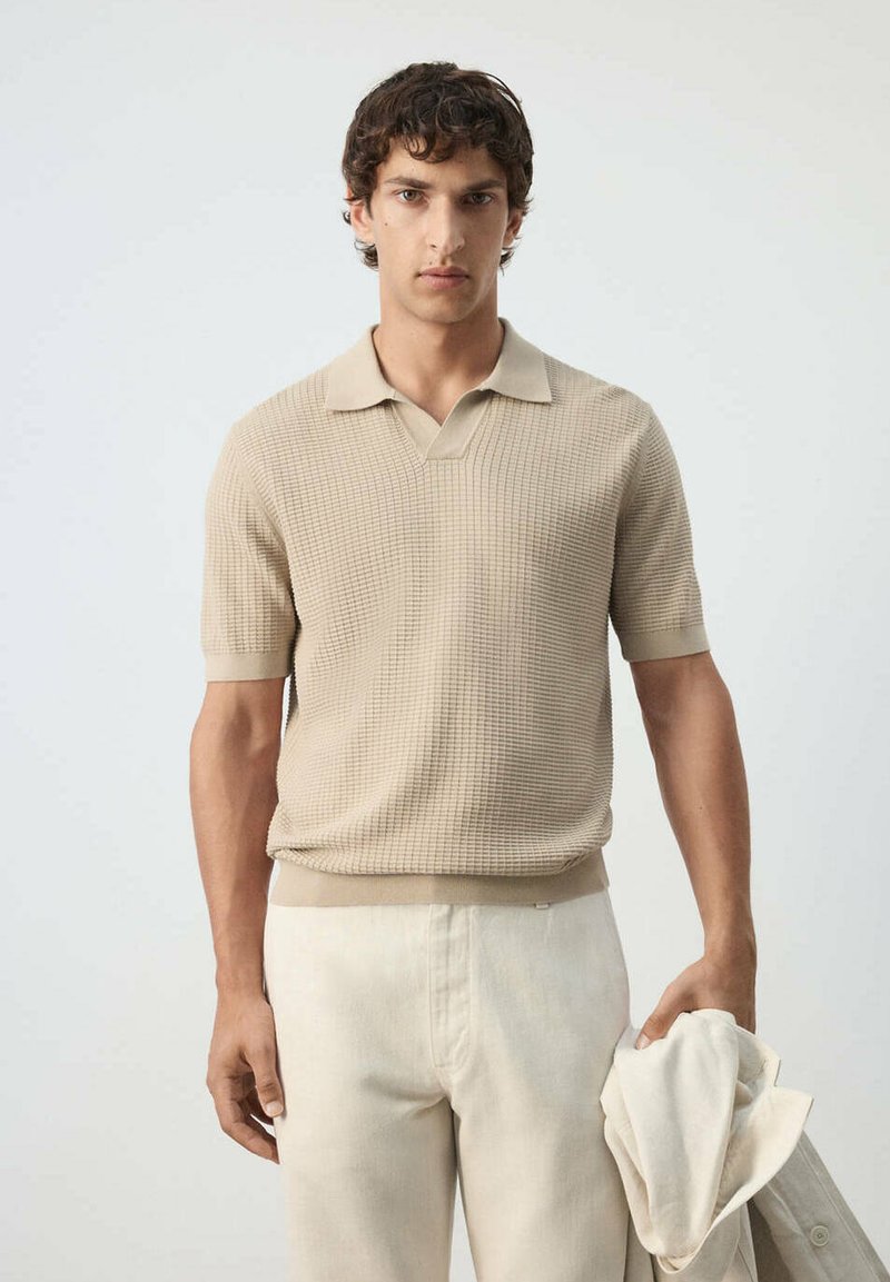 Jeune homme portant un polo beige texturé à manches courtes et un pantalon crème, tenant une veste de couleur claire dans sa main droite.