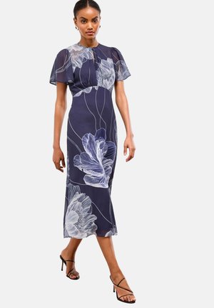 Ženska v mornarsko modri midi obleki z velikimi belimi cvetličnimi vzorci in prozornimi kratkimi rokavi, ki hodi v črnih sandalih s trakovi in peto.