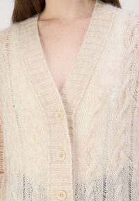 Cardigan tricotat de culoare bej deschis, având o gât de tip V adânc, cu modele texturate de cable și patru nasturi. Textură moale și pufoasă cu detalii ribbate.