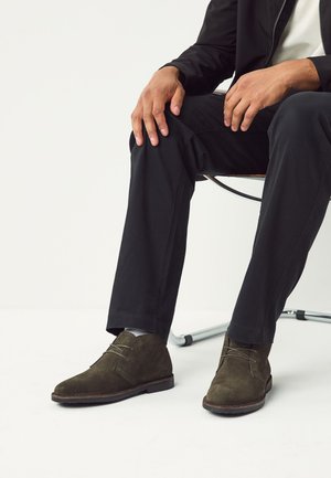 Mann sitzt auf einem Stuhl, trägt schwarze Hose, dunkelgrüne Wildleder-Schnürstiefel, weiße Socken und eine schwarze langärmelige Jacke.