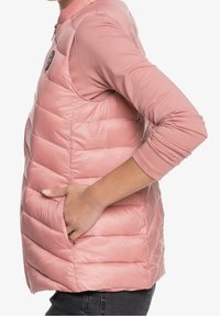 Gewatteerd roze vest met een hoge kraag, zijzak en een doorgestikt patroon. Gelaagd over een lichtroze top met lange mouwen, met een aansluitend ontwerp.