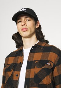 Brun och svart rutigt flanellskjorta med två bröstfickor, i kombination med en vit t-shirt och en svart keps med en vit logotyp.