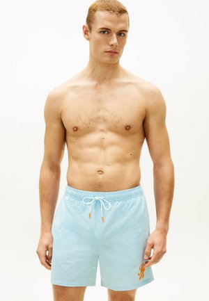 Uomo senza maglietta che indossa pantaloncini da bagno azzurri chiaro con coulisse bianca e logo arancione, in piedi contro uno sfondo bianco semplice.