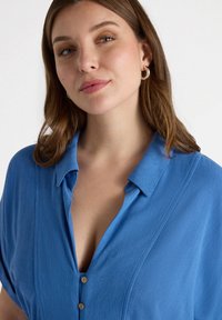 Camicia azzurra con colletto e bottoni, realizzata in un tessuto testurizzato e con maniche corte. Due bottoni in legno al collo, design a vestibilità rilassata.