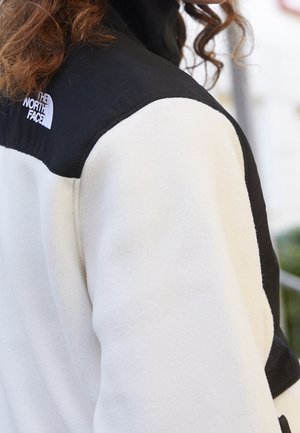 Osoba nosząca czarno-białą polarową kurtkę z logo The North Face na górnej części pleców, pokazująca kręcone, brązowe włosy.