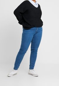 Suéter negro de punto con escote en V, combinado con jeans ajustados azules y zapatillas blancas. El suéter tiene un corte holgado y una textura acanalada.