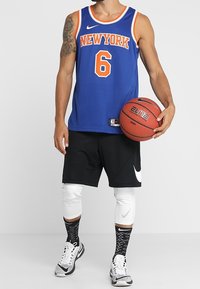 Blå baskettröja med orange detaljer och texten "NEW YORK", i kombination med svarta shorts, vita vadöverdrag och sneakers i svart/vitt.