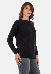 Donna con lunghi capelli castani che indossa un maglione nero a maniche lunghe e jeans grigi, in piedi con una mano sul fianco contro uno sfondo bianco.