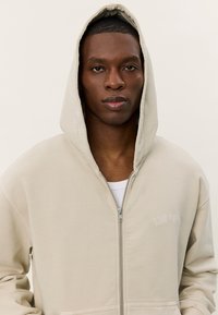 Beige zip-up hoodie with a lined hood, textured fabric, and subtle embroidered branding on the chest. Features a front zipper and side pockets.