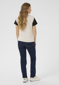 Lichtbeige mouwloze trui met een geribbelde zoom, gedragen over een zwart T-shirt, gecombineerd met donkerblauwe jeans en witte sneakers.