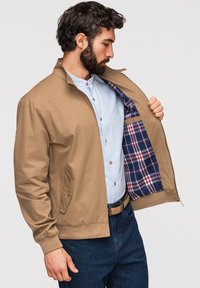 Homme portant une veste beige avec une doublure à carreaux bleu, rouge et blanc ouvre sa veste pour montrer l'intérieur par-dessus une chemise à rayures bleu clair et un jean foncé.