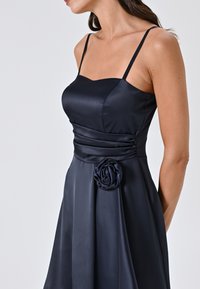 Robe en satin bleu marine avec un corsage ajusté et des bretelles spaghetti, présentant une taille froncée et une accent en rose en tissu sur la hanche.