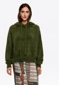 Sudadera con capucha verde y esponjosa, con cordones, combinada con una falda de cuadros estampada que presenta varios colores y texturas.