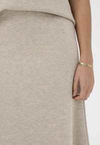 Bēšas krāsas, teksturēta auduma drapējumi maigos krokos. Roka atbalstās uz apakšmalas, demonstrējot plānu zelta aproci.