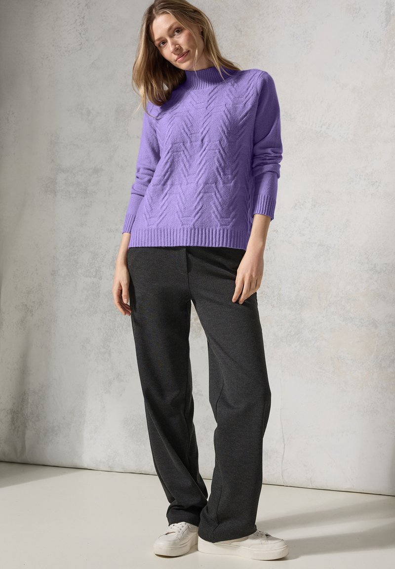 Pull en maille violet avec un motif en chevrons surélevé, col rond, manches à poignets côtelés, associé à un pantalon gris foncé et des baskets blanches sur un fond clair.