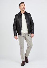 Zwarte leren jas met een staande kraag, voorzakken en een ritssluiting, gedragen over een lichtgekleurde trui en grijze broek.