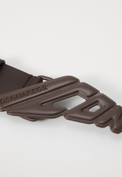 Brown rubber logo design, featuring embossed lettering. Smooth texture with cut-out shapes, attached to a brown fabric strap.