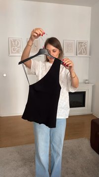 Canotta nera a coste con una spalla, orlo leggermente svasato, appesa a un gancio. Sullo sfondo ci sono pareti neutre e un pavimento in legno.