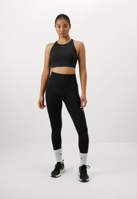 Svart sports-bh og høytlivte leggings, begge i et glatt materiale. antrekket har en liten logo på venstre hofte og sko med detaljer.
