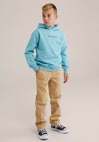 Felpa azzurra con logo frontale, abbinata a pantaloni beige e sneakers nere. La felpa ha una tasca a marsupio e polsini a costine.