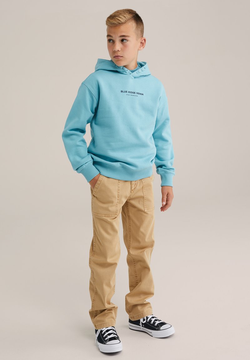 Felpa azzurra con logo frontale, abbinata a pantaloni beige e sneakers nere. La felpa ha una tasca a marsupio e polsini a costine.