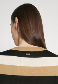 Personne aux longs cheveux bruns vue de dos portant un pull rayé beige, blanc et noir avec un logo « BOSS » sur le col.