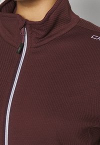 Veste zippée bourgogne en tissu texturé, dotée d'un col montant, d'une fermeture éclair contrastante blanche et violette, et d'un détail de marque sur l'épaule.