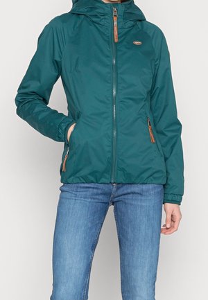 Person trägt eine türkisfarbene Kapuzenjacke mit Frontreißverschluss und Reißverschlusstaschen, kombiniert mit blauen Jeans, stehend vor einem einfarbigen Hintergrund.