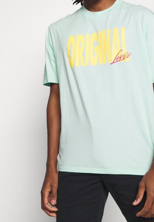 Persoon draagt een lichtgroen T-shirt met grote gele tekst "ORIGINAL" en kleinere rode tekst "Levi's", gecombineerd met een zwarte broek.