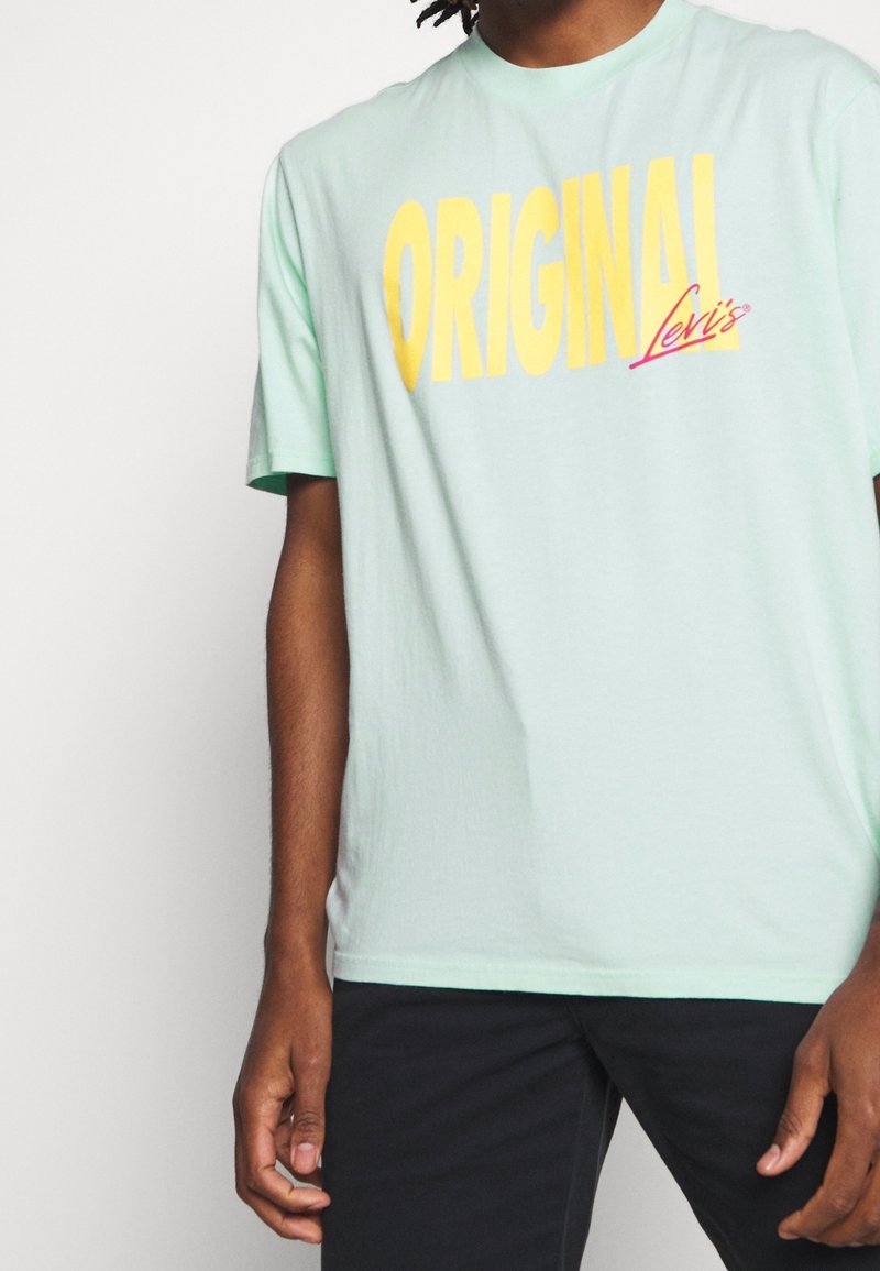 Personne portant un t-shirt vert clair avec un grand texte jaune "ORIGINAL" et un texte rouge plus petit "Levi's", associé à un pantalon noir.