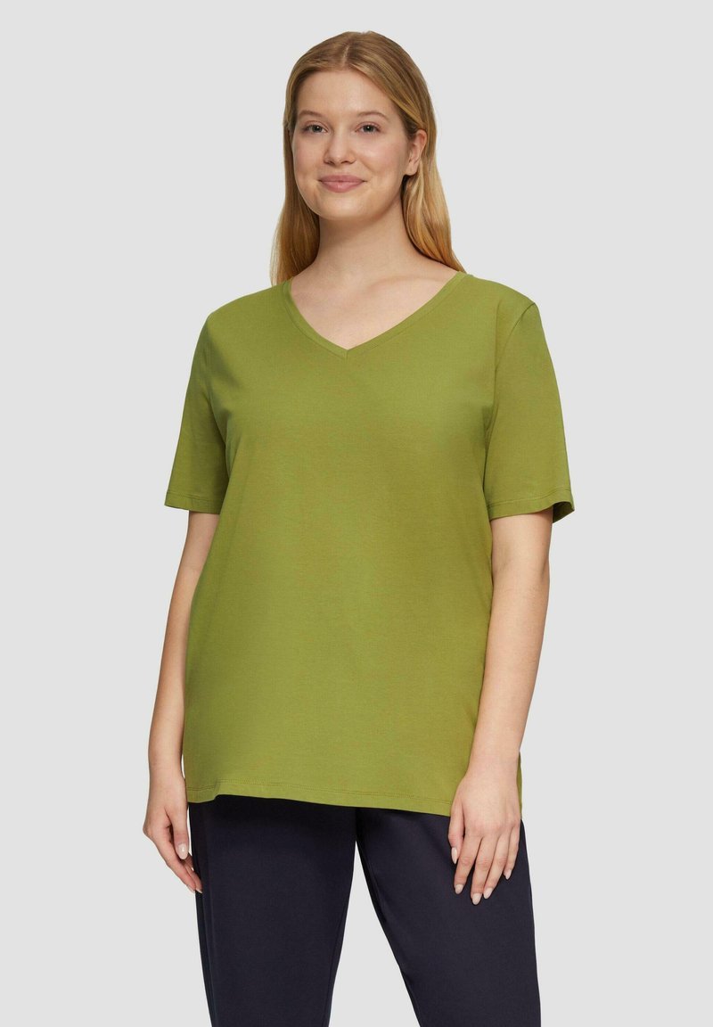Jonge vrouw die staat, gekleed in een groene korte mouwen V-hals shirt en donkere broek, neutrale achtergrond, lichte glimlach, ontspannen houding.