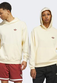Cream-colored hoodies featuring a front pocket and ribbed cuffs, adorned with a small red logo, worn with contrasting shorts.