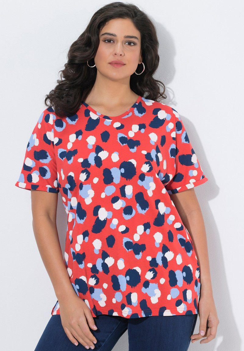 Mujer vestida con una blusa roja de manga corta con manchas abstractas en azul marino, azul claro y blanco, combinada con jeans azul oscuro y pendientes de aro plateados.