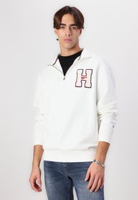 Felpa bianca con zip e logo "H" ricamato in rosso e blu. Presenta un colletto alto, polsini a costine e una texture liscia. Abbinata a jeans blu.