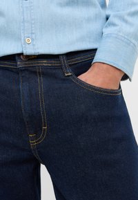 Granatowe dżinsy z kontrastowym żółtym przeszyciem, klasyczny pięciokieszeniowy fason oraz zapięcie na guziki, w połączeniu z jasnoniebieską koszulą.