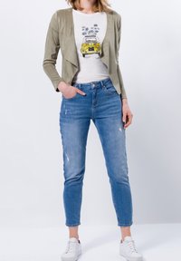 Oliven grøn cardigan med åben front, hvid T-shirt med et grafisk motiv af en gul vintage bil, og lyseblå slidte skinny jeans. Hvide sneakers.