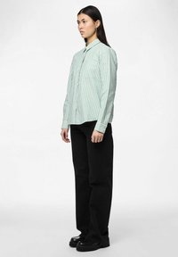 Lichtgroen button-up overhemd met witte verticale strepen, lange mouwen en een borstzak, gecombineerd met zwarte wijde broek en chunky schoenen.